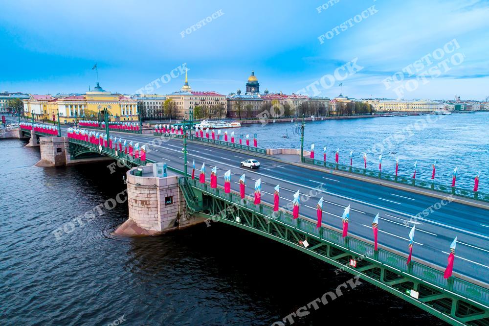 На фотографии изображён Дворцовый мост в Санкт-Петербурге, украшенный флагами в честь праздничного мероприятия. Этот знаковый мост соединяет центр города с Васильевским островом, проходя над рекой Невой. На заднем плане видны исторические здания, включая Исаакиевский собор и Адмиралтейство, которые подчёркивают величие и культурное наследие Санкт-Петербурга. Дворцовый мост является одним из самых узнаваемых и любимых мостов города, особенно впечатляющим в утреннем свете.